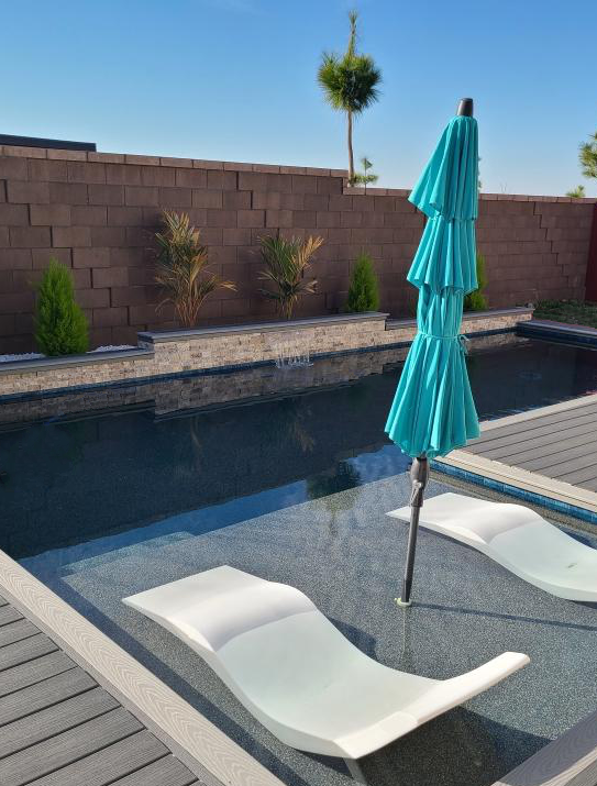 Rectangular shaped pool with a bumped out Cabo shelf with an umbrella and chaise lounges plus a raised wall with a sheer descent along with Trex decking and coping.
